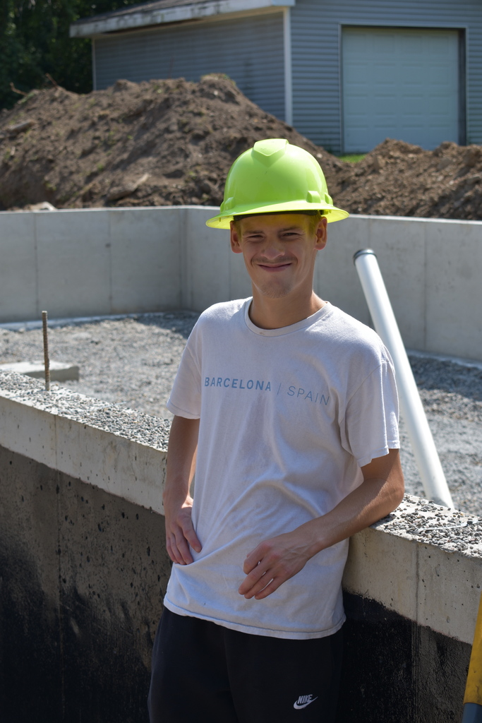 A student leaning against the foundation and smiling at the camera while wearing a hardhat 