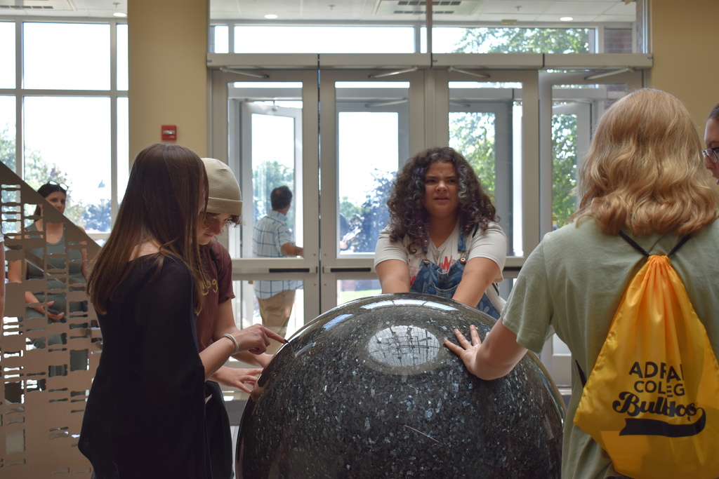 Students standing around a circular art structure with running water. Each student is touching the water as they stand around it and talk to each other.