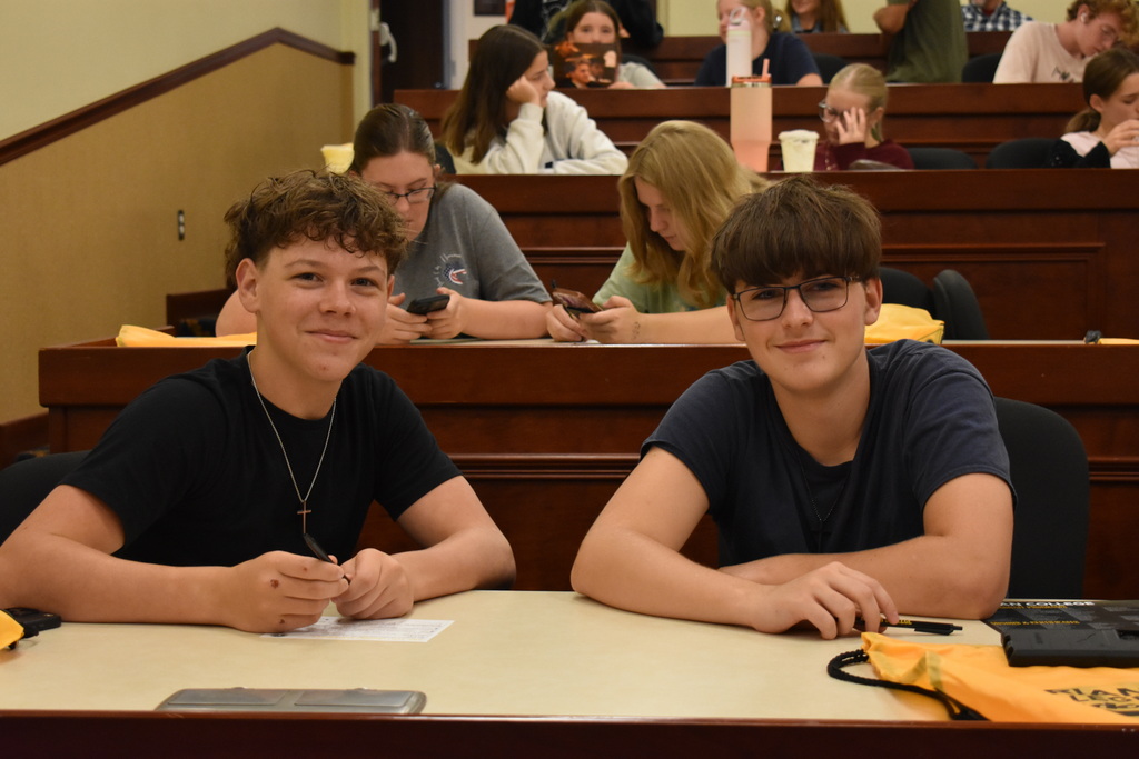 Two students looking at the camera and smiling while in an auditorium with students seated behind them at Adrian College