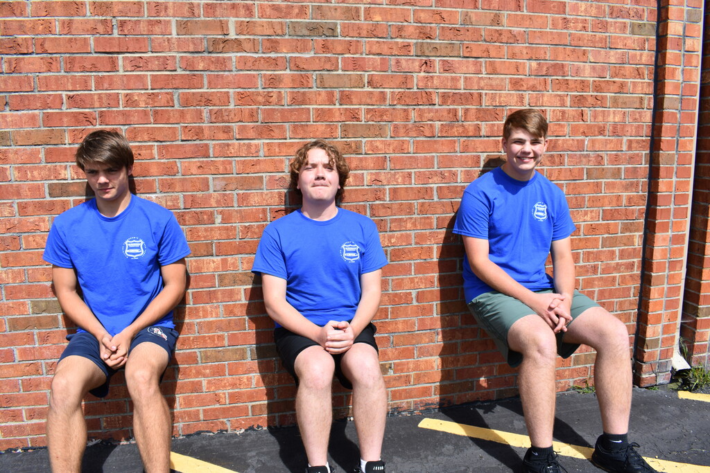 Three students holding a wall sit outside against a brick wall
