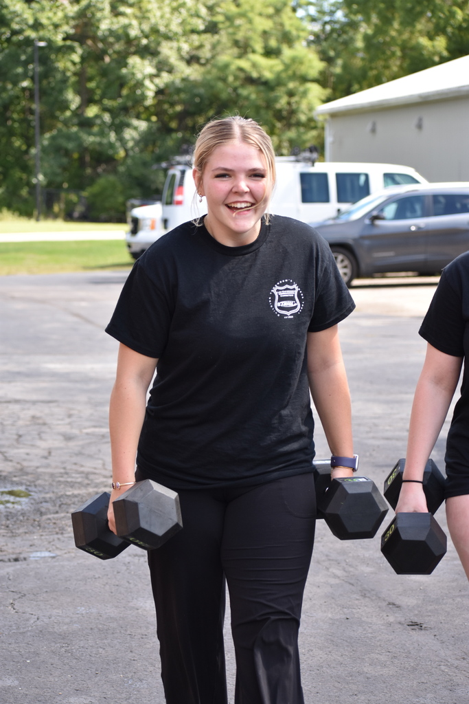 A student smiling as she carries dumbbells for a farmer's walk outside