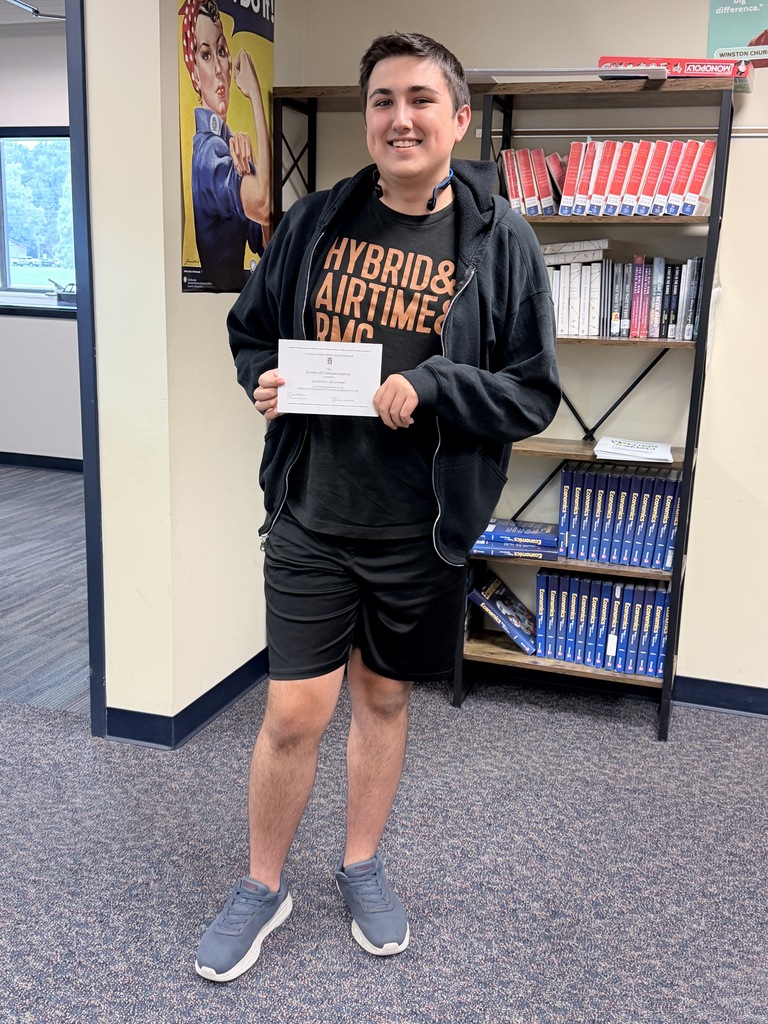 Student Tony Grudnoski holding a certificate and smiling at the camera in front of a book shelf in a classroom setting