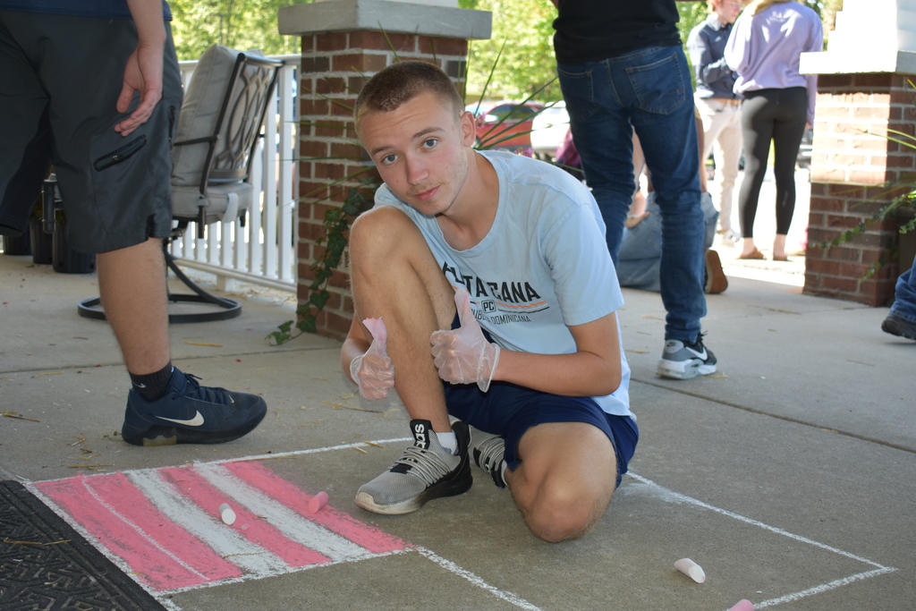 A male student kneeing next to a flag he is creating using chalk and giving the thumbs to the camera
