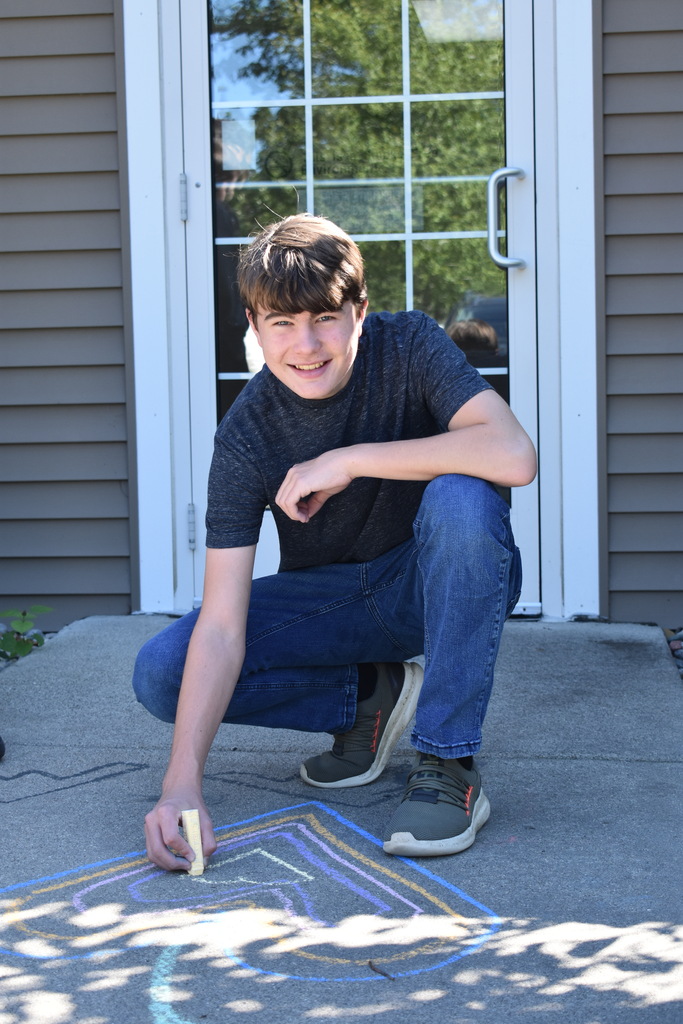 A male student using sidewalk chalk to create a design. He is smiling at the camera while kneeling down on the sidewalk.