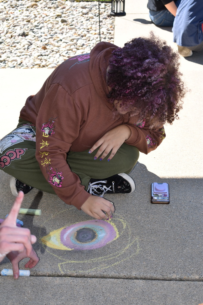 A student working on a design of an eye using sidewalk chalk while seated on the ground