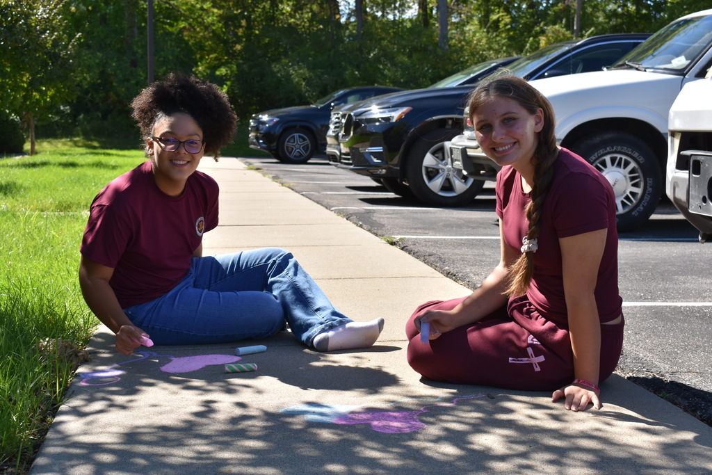 Two students looking at the camera and smiling while working on artwork using chalk on a sidewalk. Both students are seated.