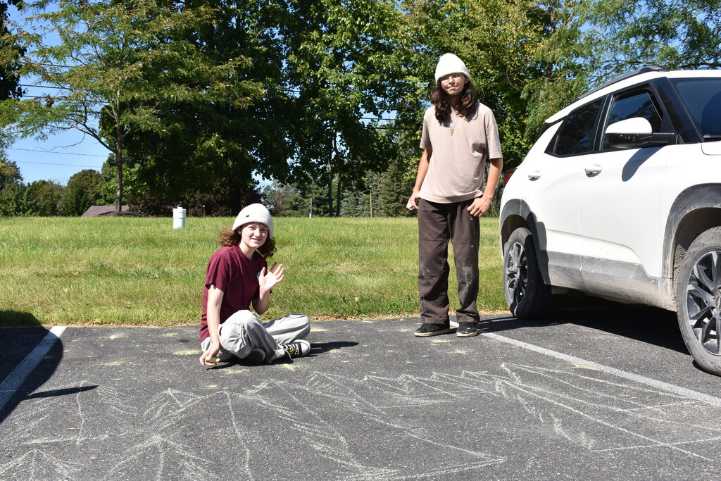 Two students, one sitting and one standing, in a parking lot spot over their artwork. The seated student is waving at the camera and both are looking at the camera and smiling.