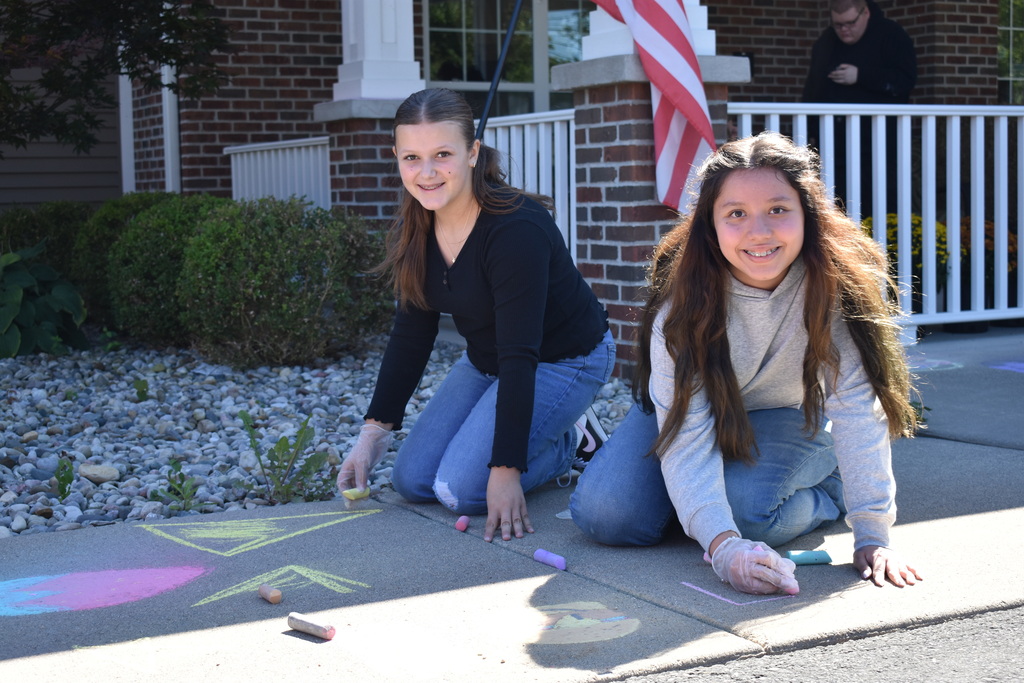 Two students outside a senior living facility using sidewalk chalk on a walkway while both looking at the camera and smiling.
