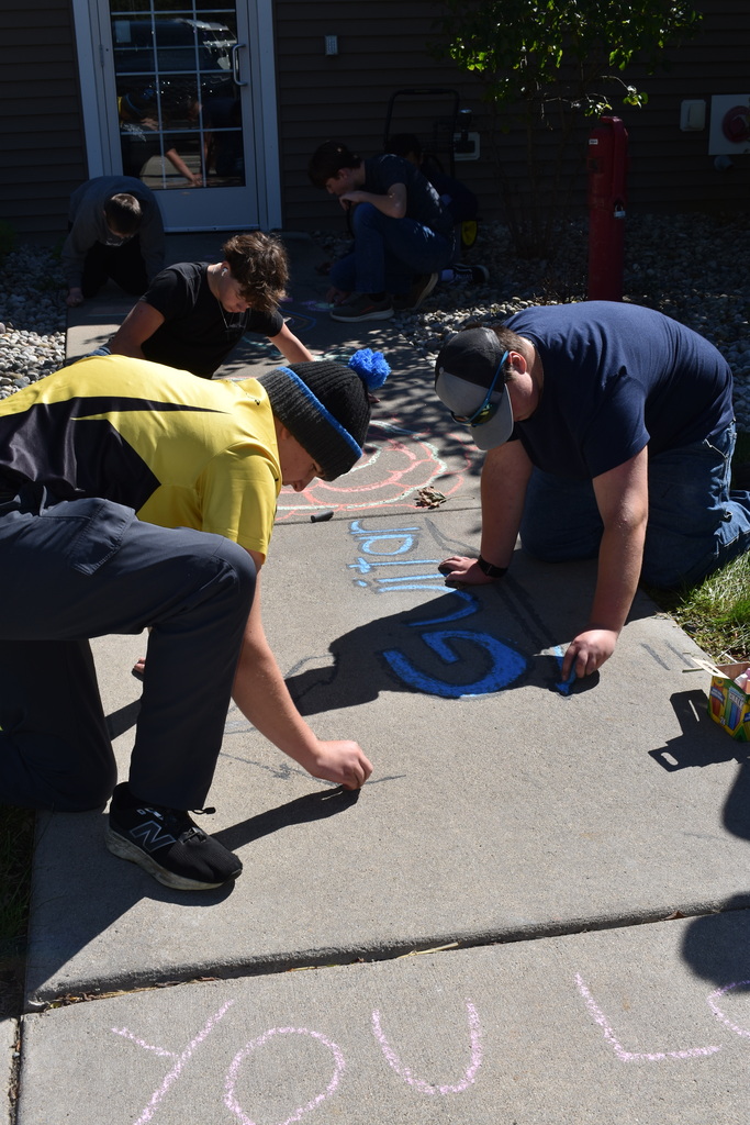 Five male students kneeling on the ground doing sidewalk chalk art on a sidewalk outside a senior center.