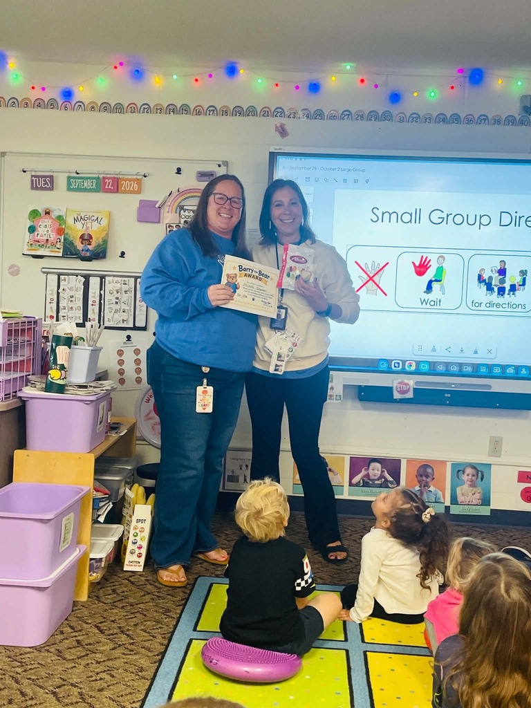 Michelle Bowman and Kari Truax smile together at the front of a classroom as Michelle holds her Barry the Bear Award certificate. Students sit on the carpet in front of them during the presentation.