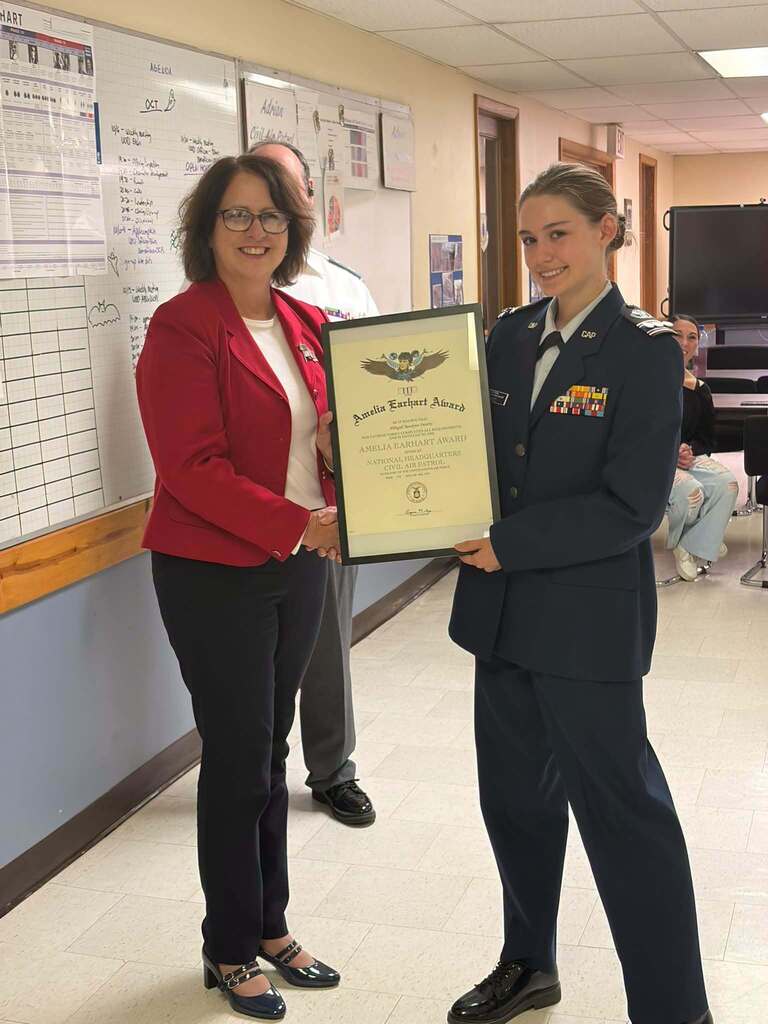 JC/LISD Academy student Abbigail Swartz, dressed in her Civil Air Patrol uniform, smiles as she receives the prestigious Amelia Earhart Award. She shakes hands with a woman in a red blazer while they hold the framed award certificate together.