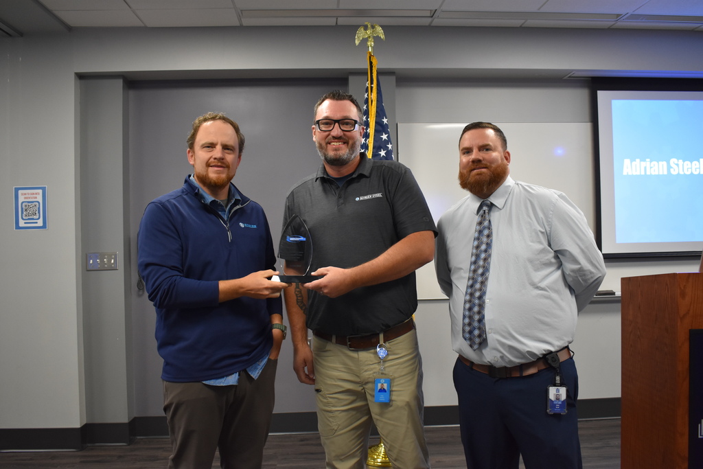 Business partner of the year award being given to Adrian Steel. Two employees from Adrian Steel standing with Assistant Principal Jake Graf with an American flag behind them in a classroom setting.