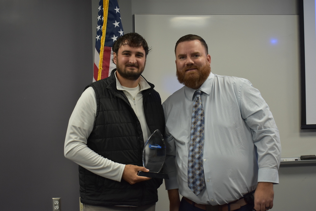 An alumni award being given to Matt from Assistant Principal Jake Graf. The two posing together in a classroom setting with an American flag behind them.