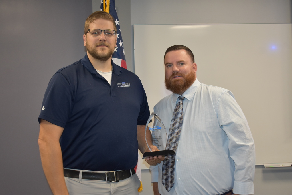 An alumni award being given to Kory from Assistant Principal Jake Graf. The two posing together in a classroom setting with an American flag behind them.