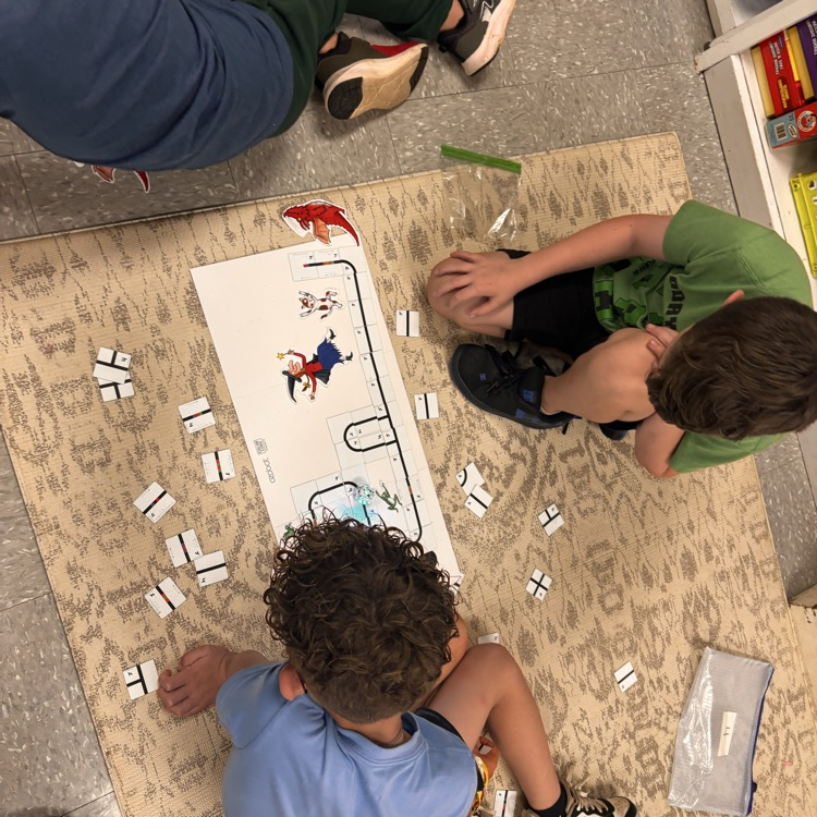 students sit on the ground, there is a white mat with magnets, an Ozobot, and book characters