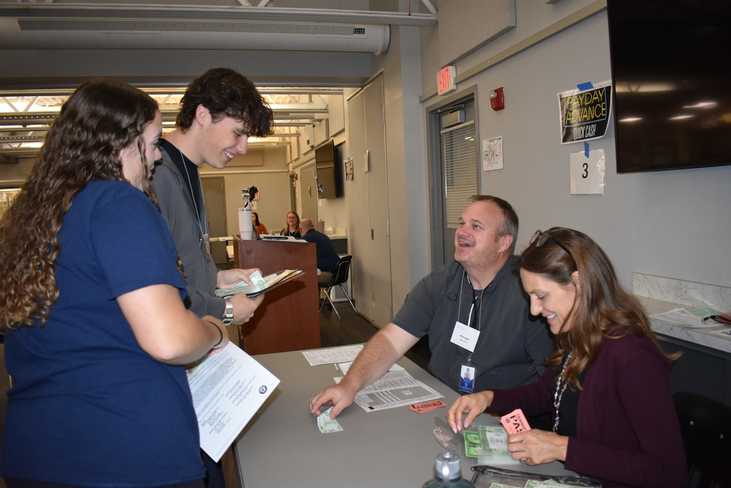 Two students interact with volunteers at a simulated payday advance station, exchanging play money and reviewing paperwork.