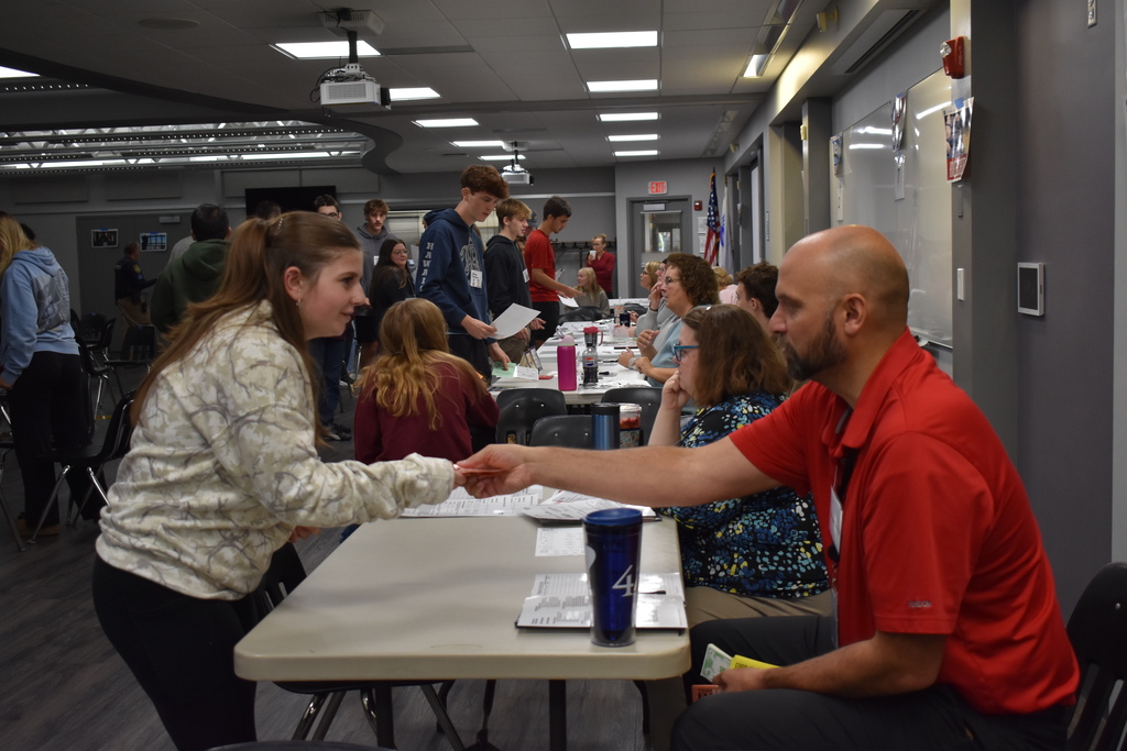 A student exchanges paperwork with a volunteer at a resource station while other students wait and participate in the simulation.