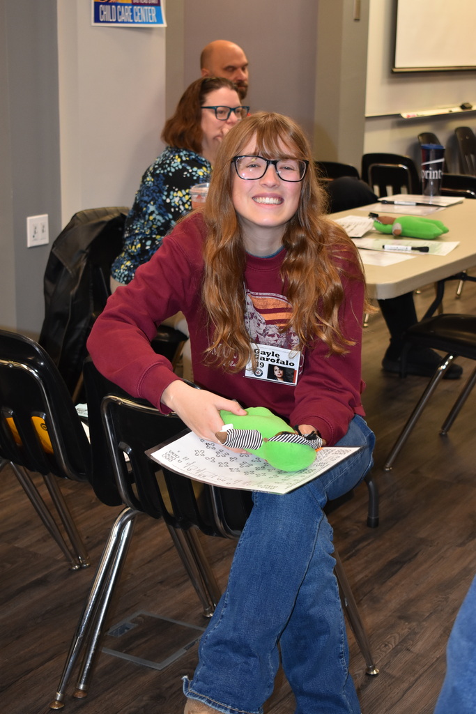A smiling student with glasses sits holding a green stuffed toy and paperwork while participating in the Poverty Simulation at the LISD TECH Center.