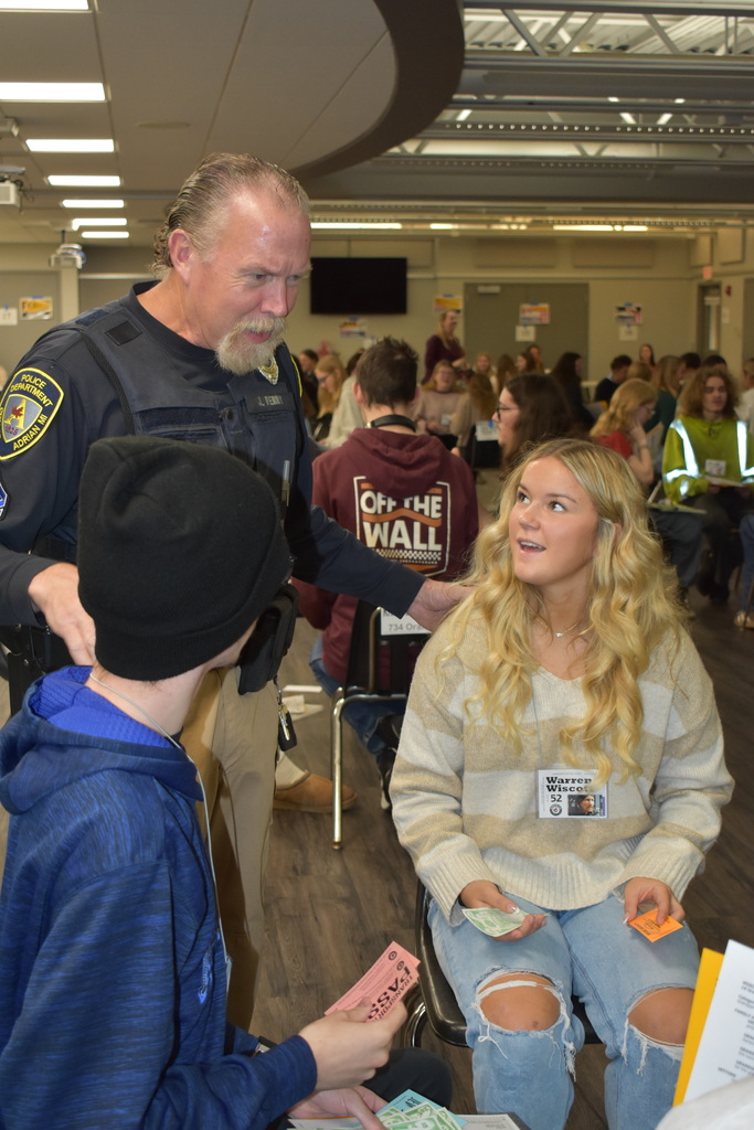 An Adrian Police Department officer speaks with two students during the Poverty Simulation as other participants move between stations.