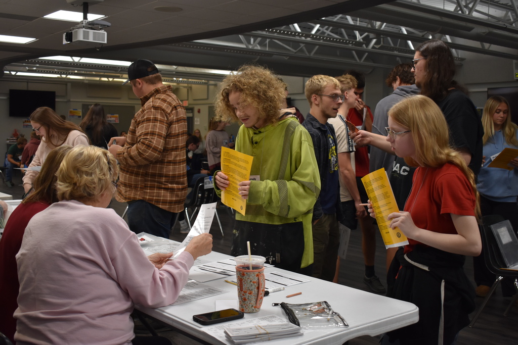 Students holding yellow information sheets interact with volunteers at a resource station during the Poverty Simulation.