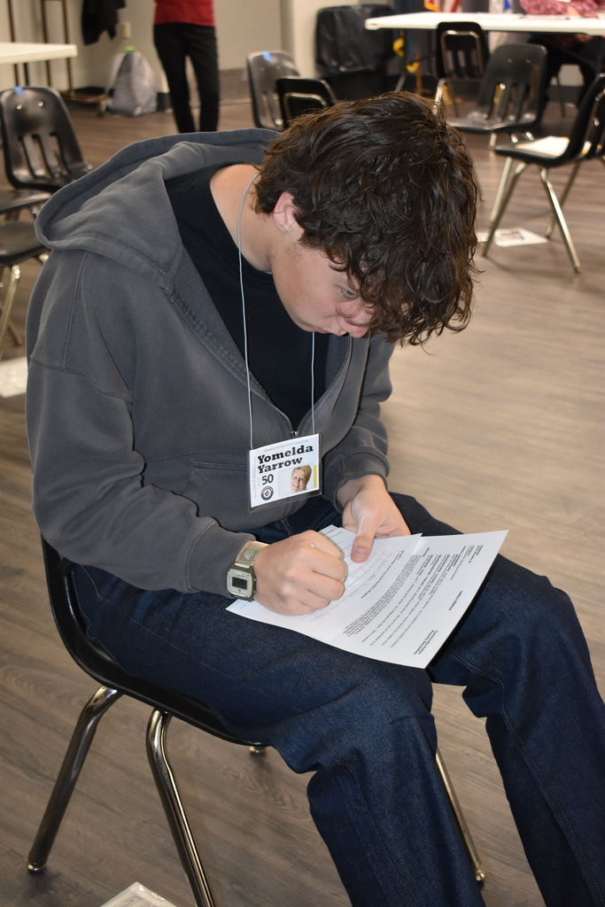 A student sits in a chair, concentrating on completing paperwork for an assigned scenario during the Poverty Simulation.