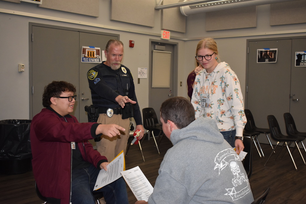 An Adrian Police Department officer speaks with a group of students holding paperwork near a simulated police department station.