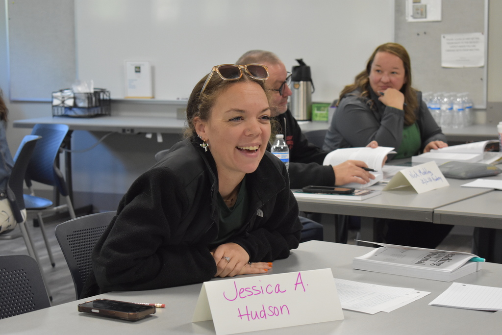 An educator smiles while participating in a literacy training discussion, with fellow educators reviewing materials at tables behind her.