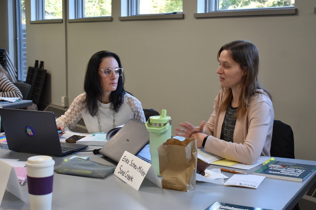 Two educators sit across from one another, discussing literacy strategies with books, notes, and a laptop spread across their table.