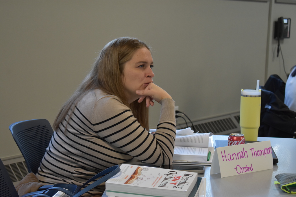 An educator from Onsted listens attentively during the Secondary Literacy Network training, with an open book, notebook, and literacy resources on the table in front of her.