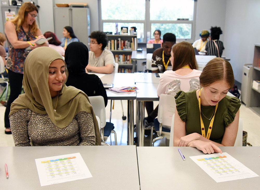 Two femal high school sophomores study at a table with classmates in the background