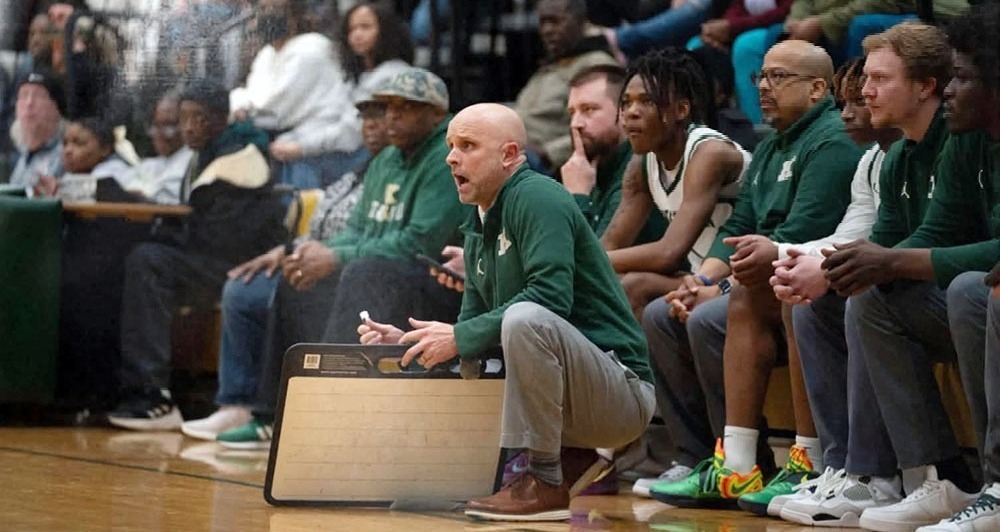 Basketball coach in green sweather kneels on the sideline, urging his team on.