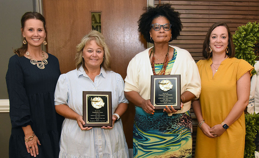 Four women pose in a line with the wo in the middle holding awards plaques.
