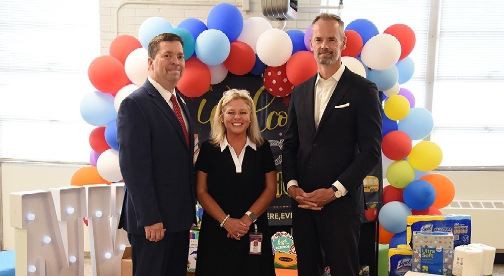 Two men in dark suits and a young woman in a blue dress pose in front of a collection of school supplies.