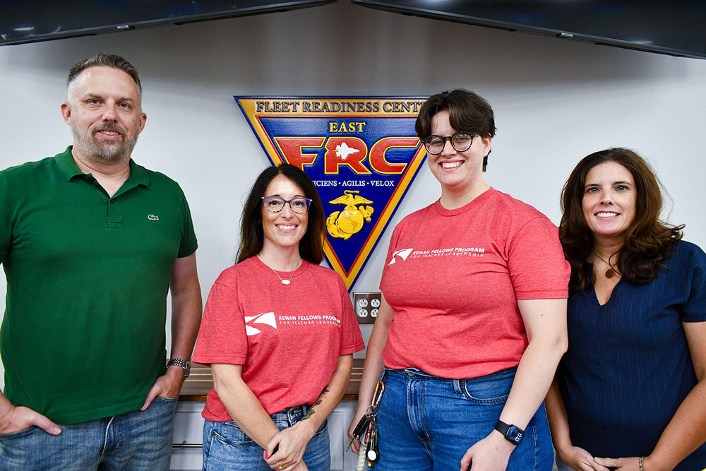 Four people, three females and a male, pose in front of an inverted triangular sign that reads Fleet Readiness Center East.