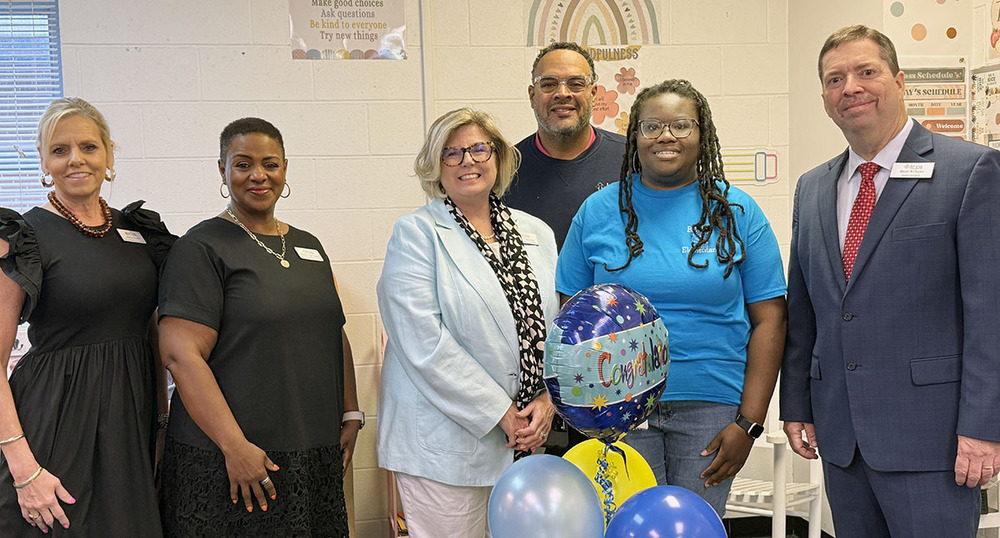 Five people pose in a line, including a woman holding balloons.