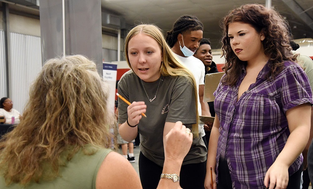 Two female high school junior face the camera as they talk to a older woman, seated at a desk with her back to the camera