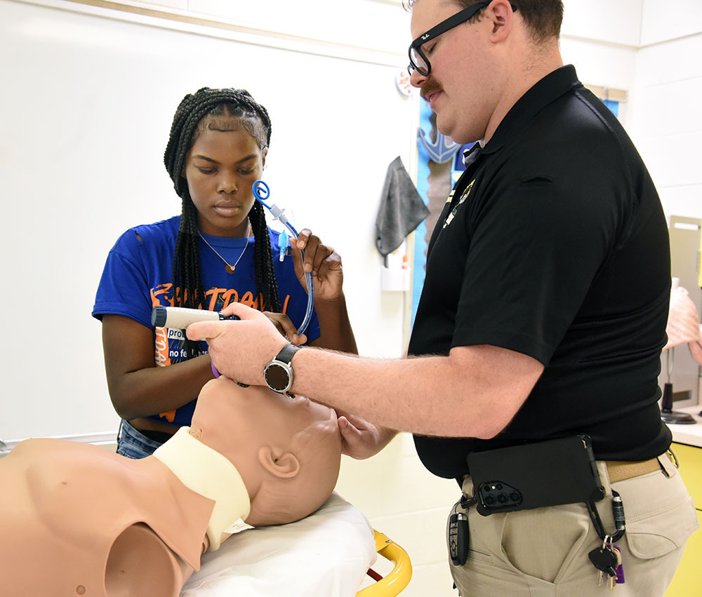 Female high school senior helps EMS training officer demonstrate a medical technique on a mannequin.