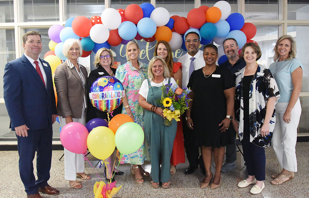 Short blonde woman holding flowers is surrounded by smiling people posing after an award presentation.