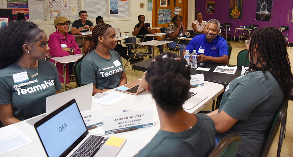 Four high school students sit at a table with their laptops talking to a businessman.
