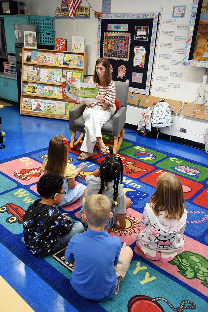 A female indergarten teachers sits in a rocking chair and reads aloud to five children sitting in front of her on the carpet.