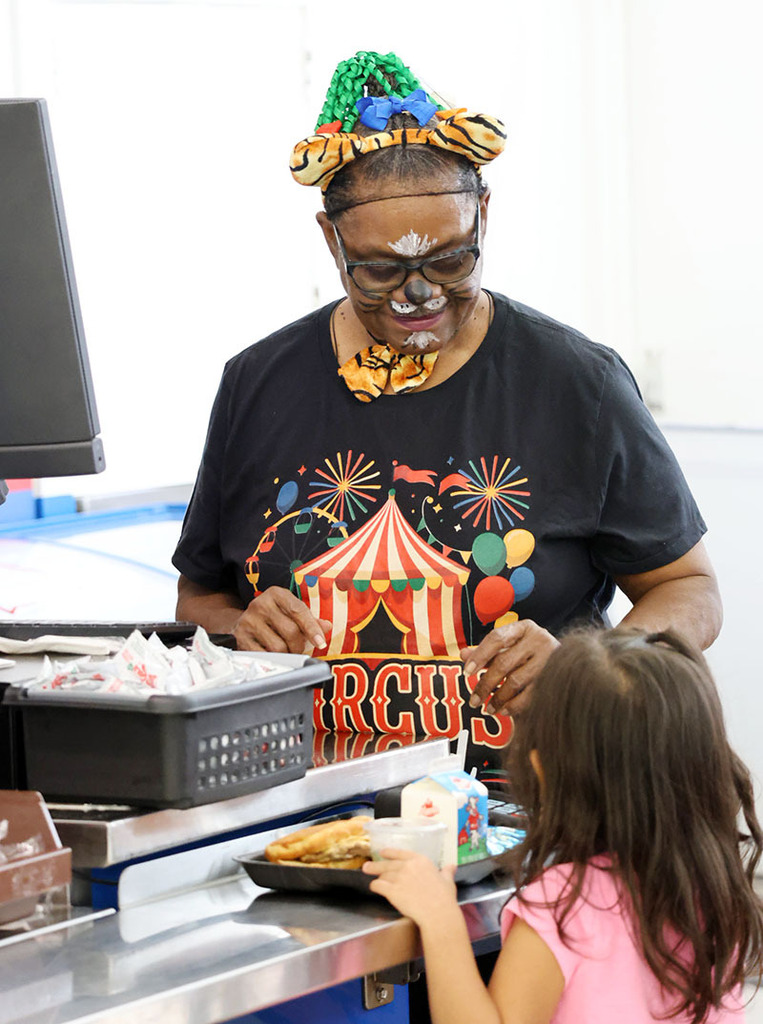 School cafeteria worker in tiger-like face paint waits on a young student.