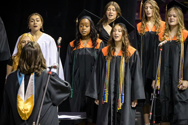 Choir sings in black and orange graduation gowns.