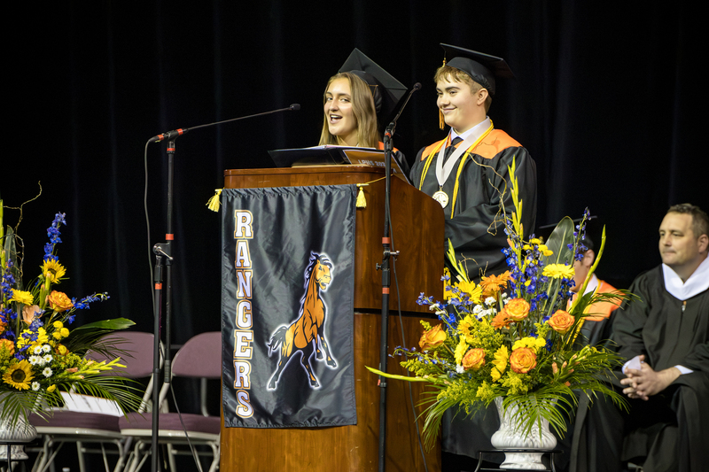 Two females speak at the podium in black gowns and caps.