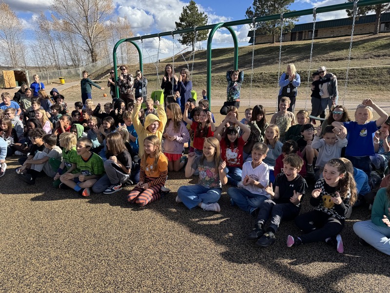 Students sit on the new playground by swings watching the ribbon cutting.