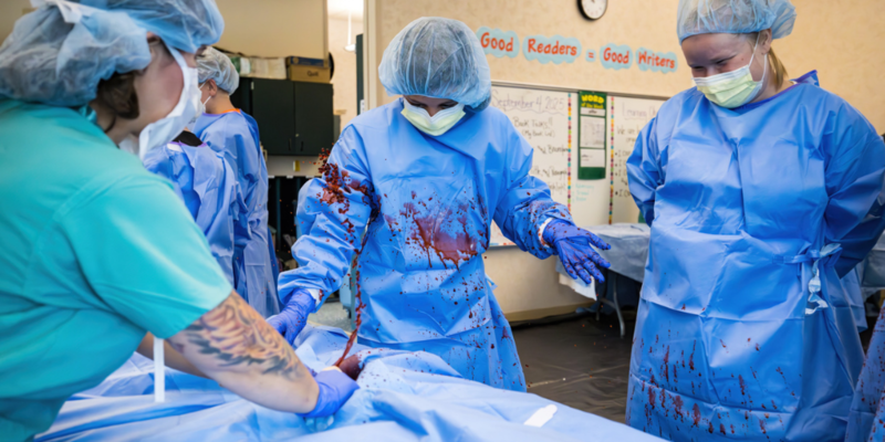 students perform surgery on medical dummy, blood spurts out onto students in scrubbs