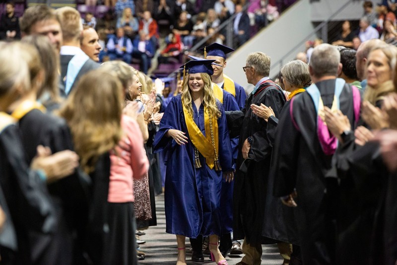 Students in graduation cap and robes walking down aisle between educators and staff in regalia