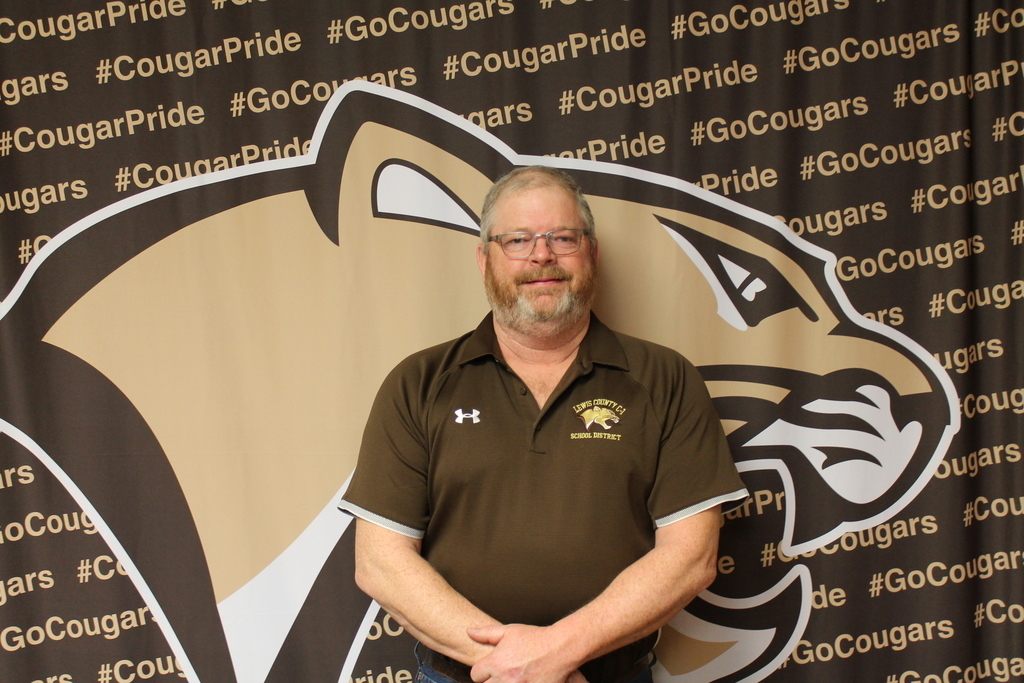 Neil Jennings, vice-president of the Lewis County C-1 Board of Education, is wearing glasses and a brown Lewis County C-1 polo shirt standing with his hands clasped in front cougar mascot backdrop filled with “#CougarPride” and “#GoCougars” text and is being recognized for his 15 years of dedicated service to the district.