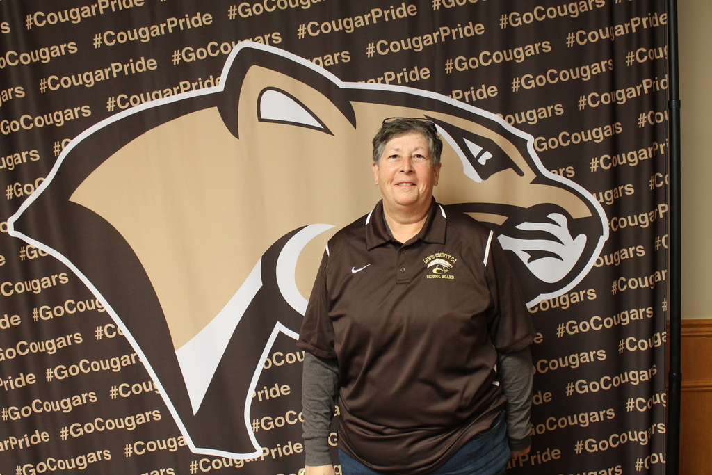 Sue Ann Gaus stands smiling in a brown school board polo shirt against a backdrop featuring a large cougar mascot logo and repeating "#CougarPride" and "#GoCougars" text.