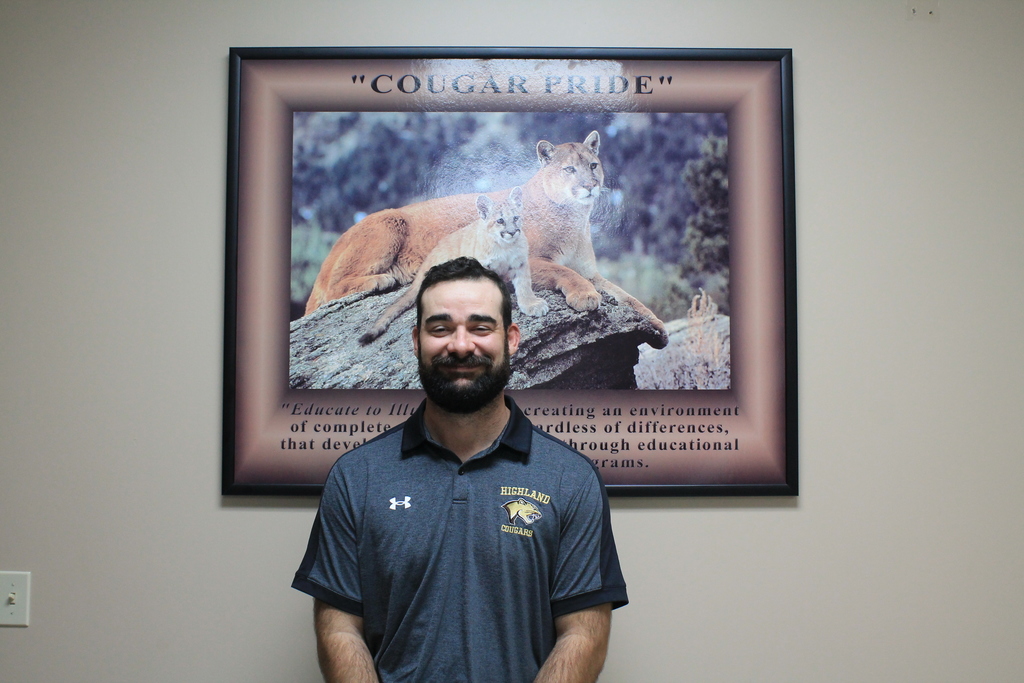 JT Gray, wearing a grey Under Armour polo shirt with "HIGHLAND COUGARS" text and a mascot, stands smiling in front of a framed poster titled "COUGAR PRIDE" featuring a photo of a cougar and cub.