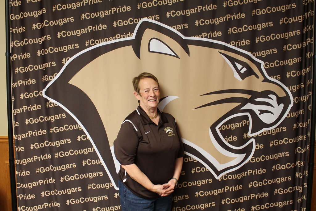 A smiling Donna Reid, wearing a brown Lewis County Board of Education polo shirt, stands against a backdrop patterned with a large gold cougar head logo and repeating #GoCougars and #CougarPride hashtags.